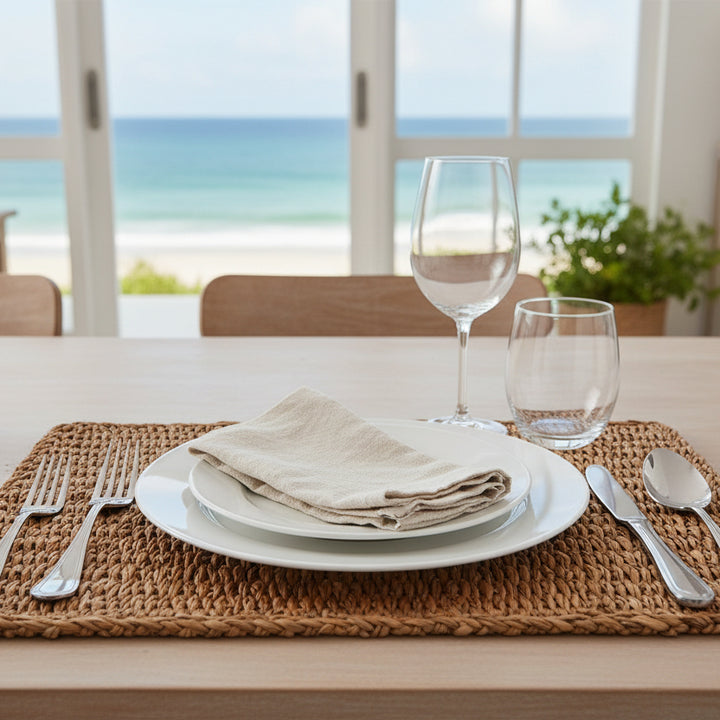 A rectangular woven seagrass placemat with a natural tan and brown color palette, featuring a thick braided border, shown against a BEACH SETTING