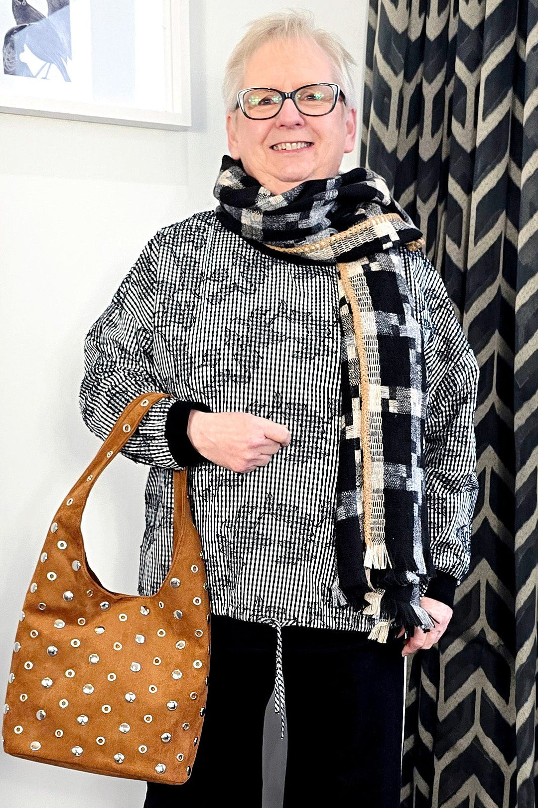 Woman standing indoors wearing a patterned scarf, gray sweater, black pants, and white shoes, with a brown patterned handbag.
