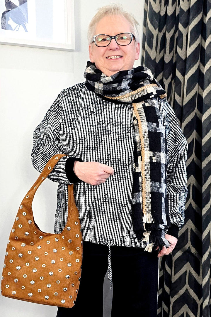 Woman standing indoors wearing a patterned scarf, gray sweater, black pants, and white shoes, with a brown patterned handbag.