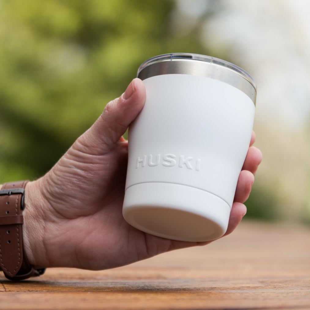 Hand holding a white Husky cup outdoors on a wooden surface with blurred greenery in the background