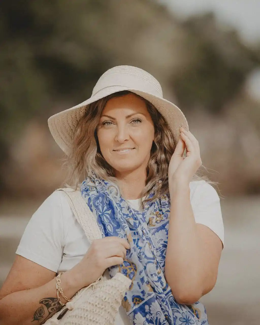 Woman wearing a wide-brimmed hat and blue scarf, standing outdoors with a blurred natural background