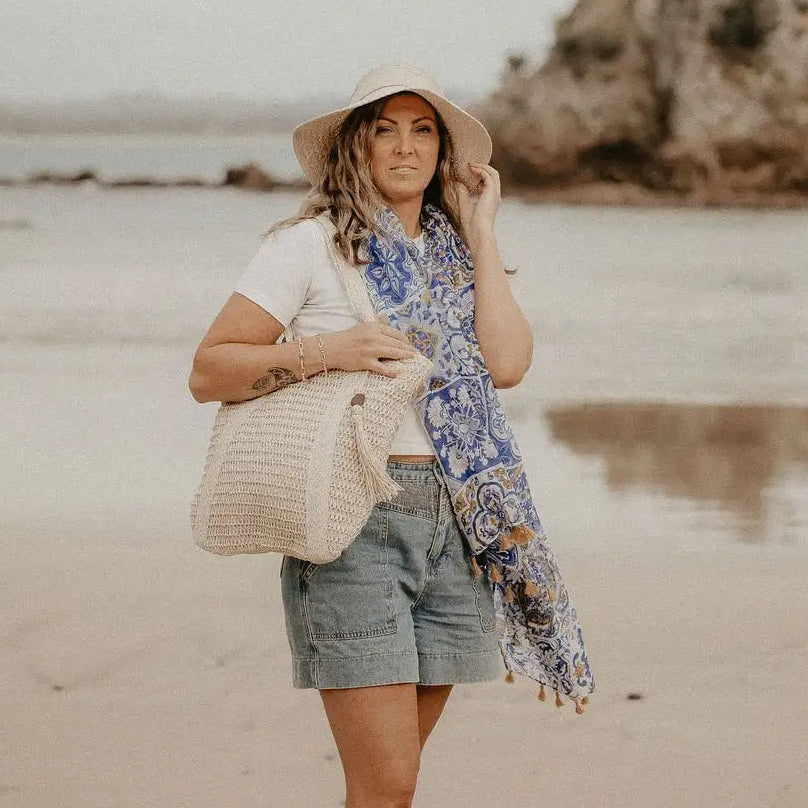 Woman on a beach wearing a hat and holding a bag, with a scenic background.