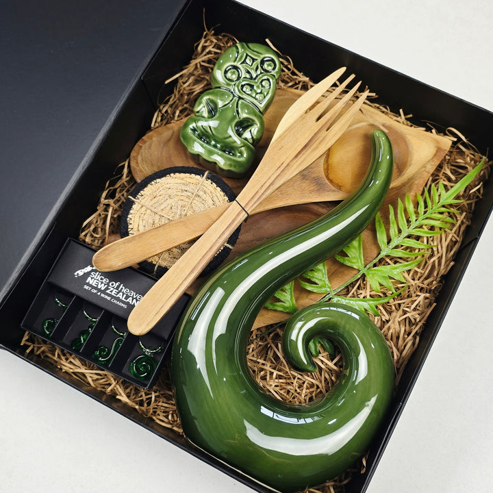 A black gift box filled with a green ceramic Maori-style carving and wooden utensils, photographed from above on a white tabletop.