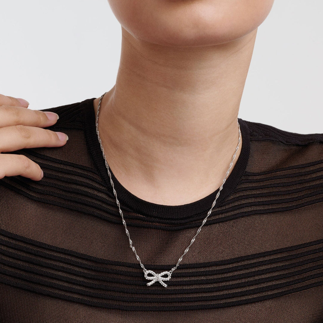 Person wearing a ted baker silver necklace with a bow pendant on a plain background