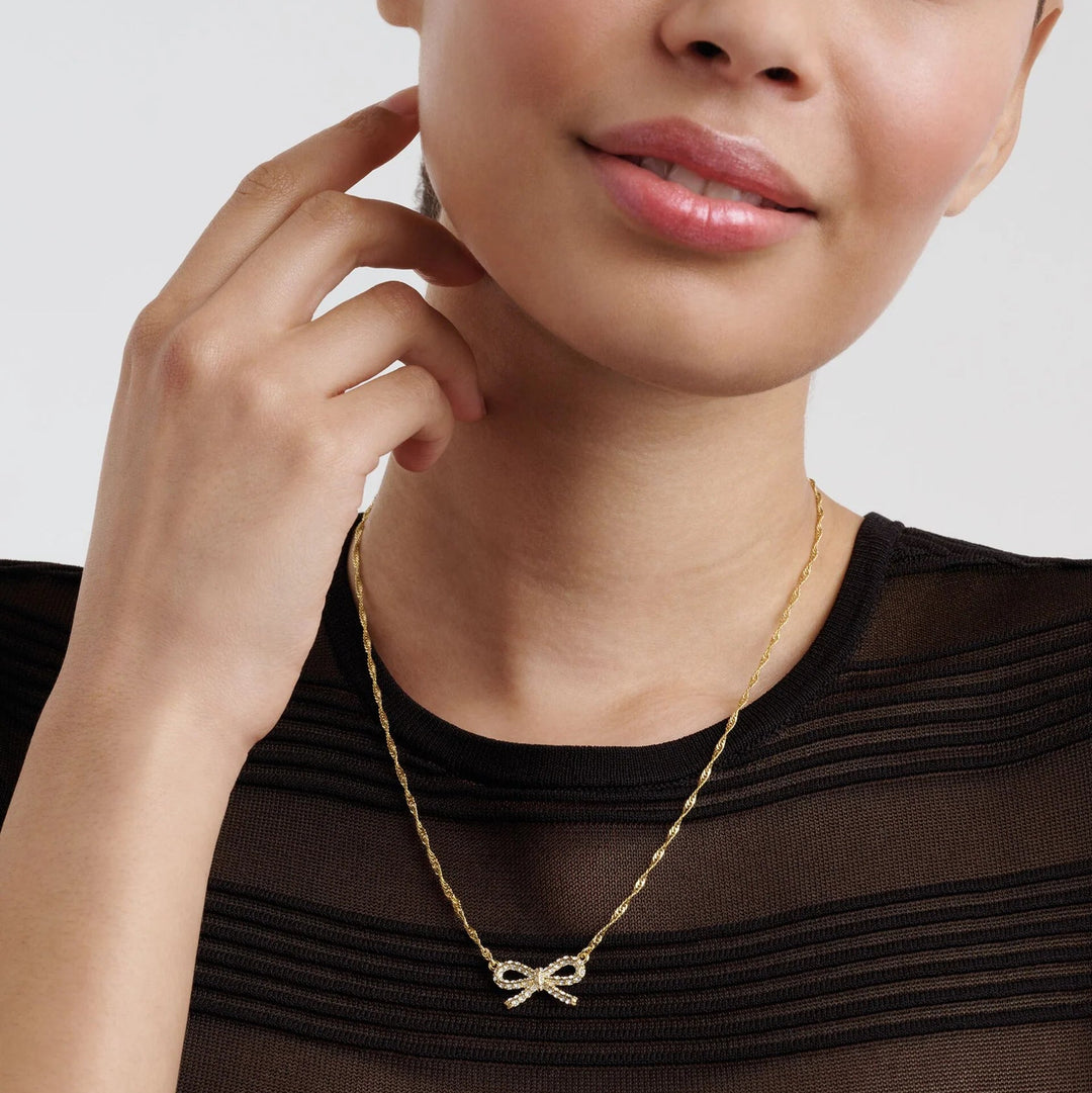 Woman wearing a gold necklace with a bow pendant against a neutral background