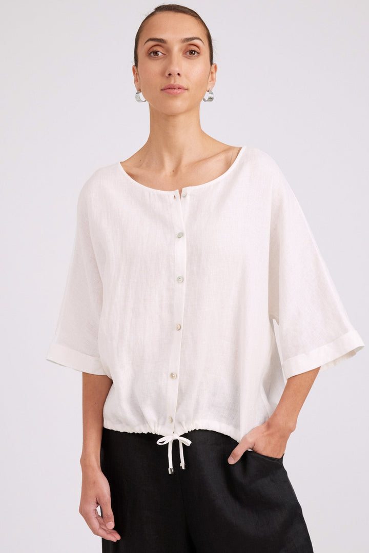 A woman wearing a loose white short-sleeve button-front blouse with a drawstring hem, paired with black pants, photographed against a light gray studio background.