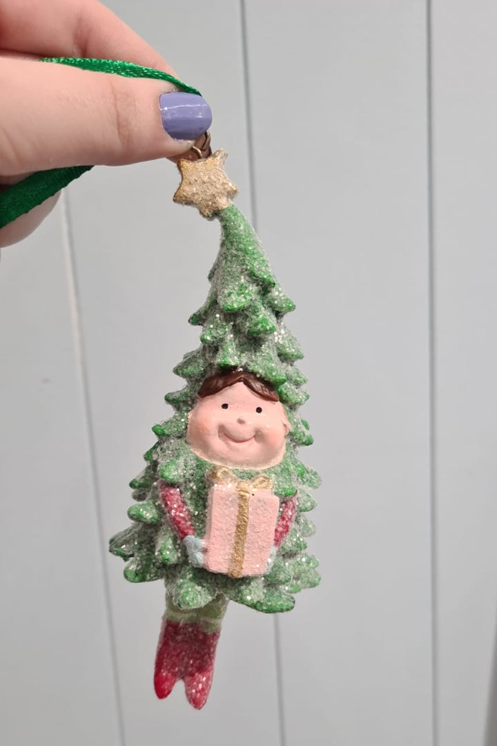 Decorative Christmas tree-shaped ornament with a smiling face and gift, held by a hand against a plain background.