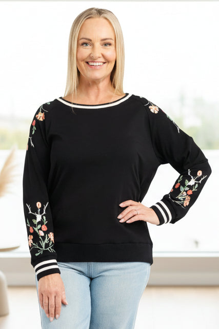 Woman wearing a black sweater with floral patterns on sleeves and light blue jeans against a white background