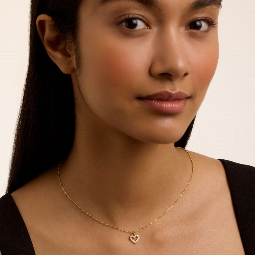 Woman wearing a ted baker gold necklace with a heart pendant on a plain background