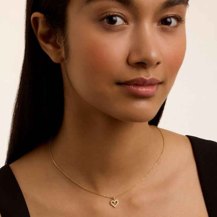 Woman wearing a ted baker gold necklace with a heart pendant on a plain background
