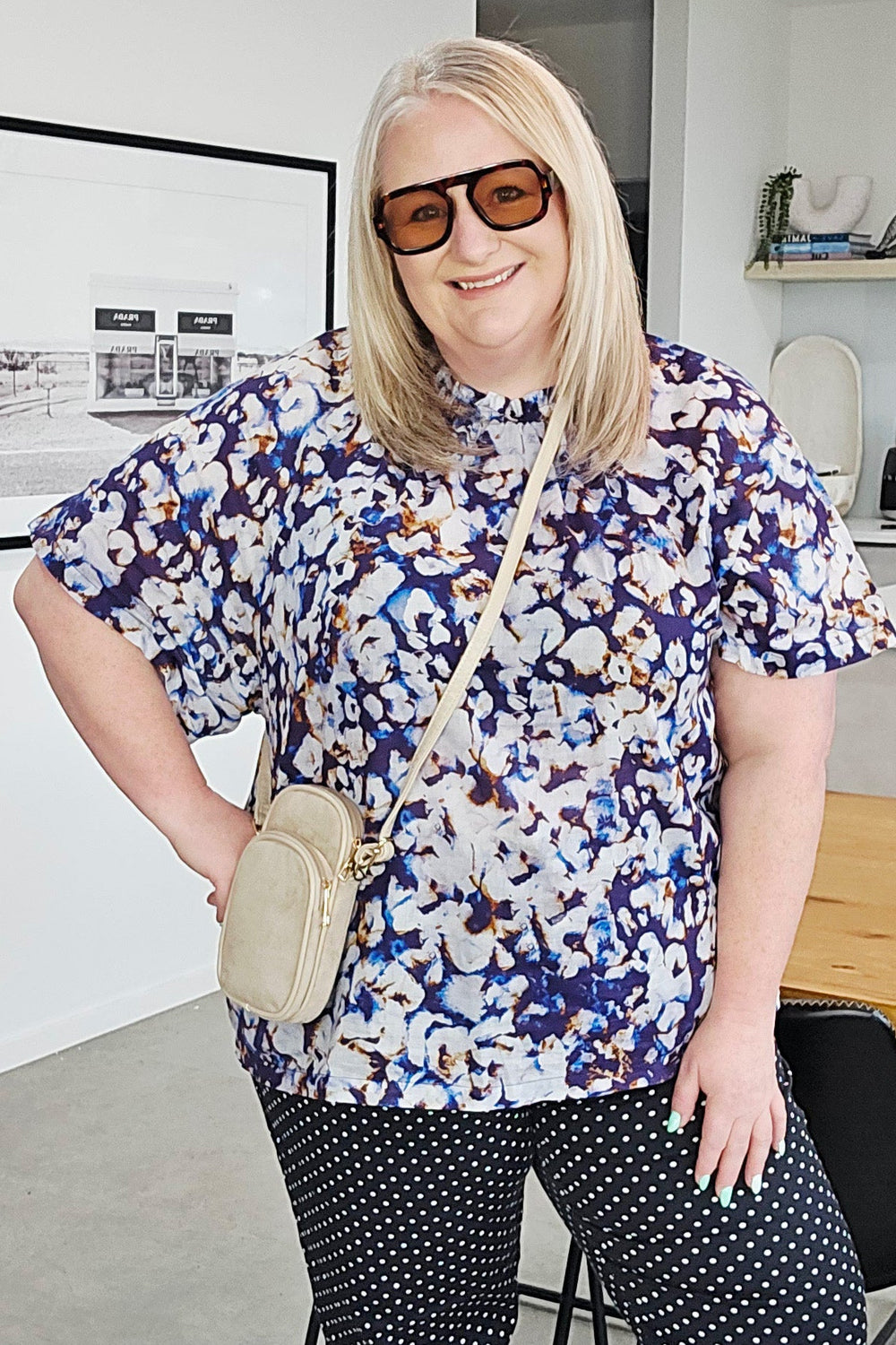 A woman modeling a short-sleeve button-up shirt with a blue, white, and black abstract pattern, paired with black and white polka dot cropped pants, white Mary Jane shoes, and a beige crossbody bag.