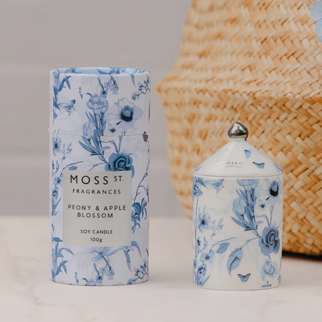 Moss St Fragrances Peony & Apple Blossom soy candle shown in its blue-and-white floral cylindrical box and matching small lidded jar, styled on a light surface with a woven basket and pale blue flowers in the background.