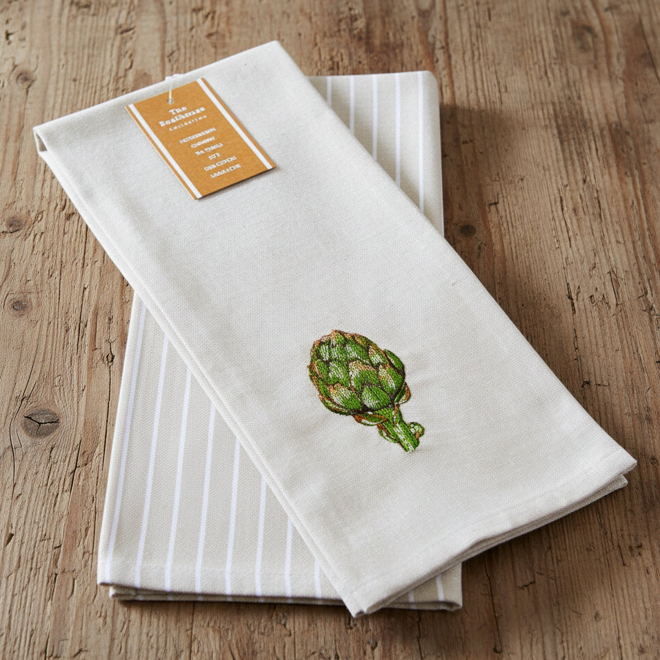 A folded beige kitchen towel with white vertical stripes and a green embroidered artichoke, featuring a tan paper label at the top.