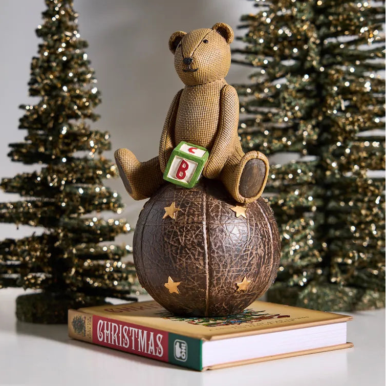 Teddy bear with a letter 'B' on a Christmas book, surrounded by decorated trees.