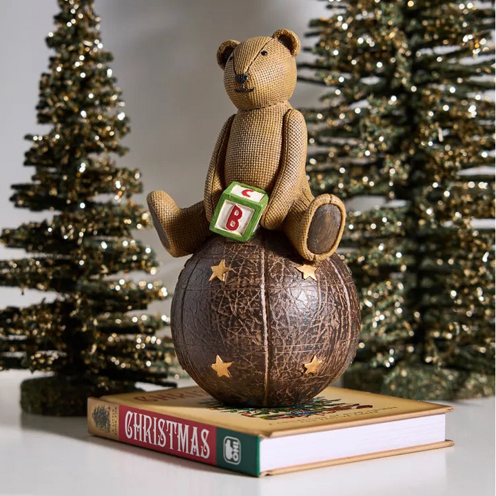 Teddy bear with a letter 'B' on a Christmas book, surrounded by decorated trees.