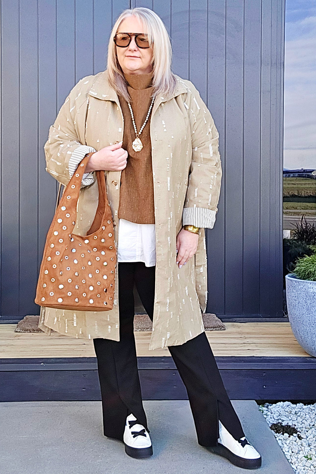 Woman in a beige coat with a patterned bag standing outdoors.