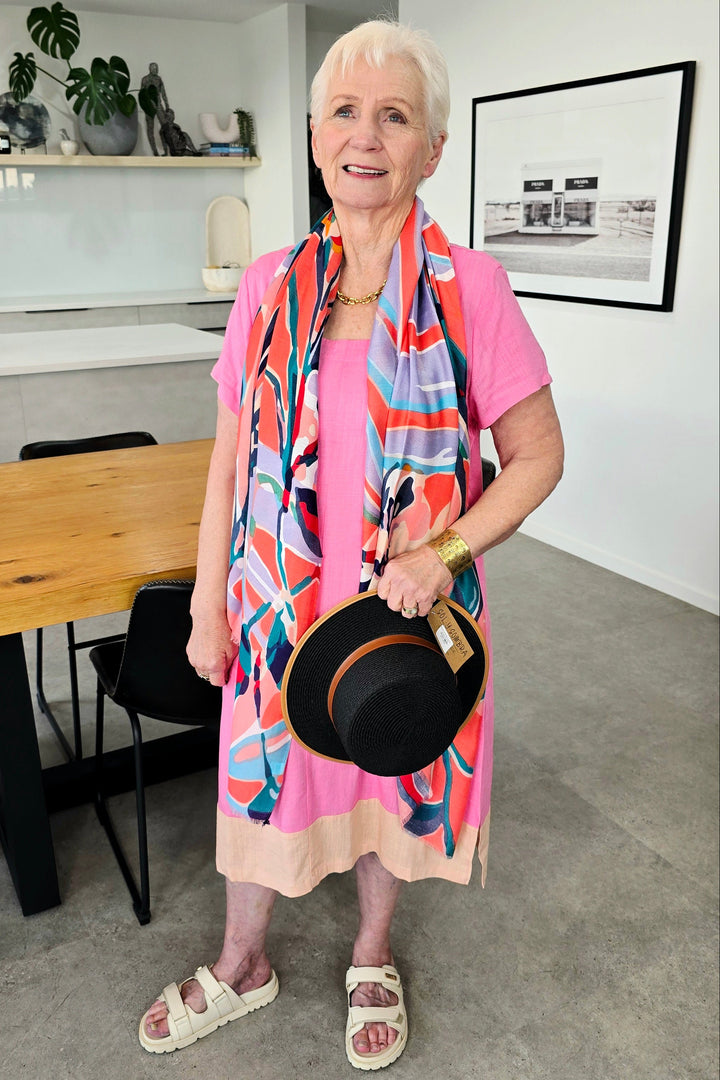 An older woman stands indoors wearing a bright pink dress with a bold multicolor abstract print scarf, holding a black hat with a brown band. The setting is a modern home interior with a wooden dining table, concrete floor, and white walls.