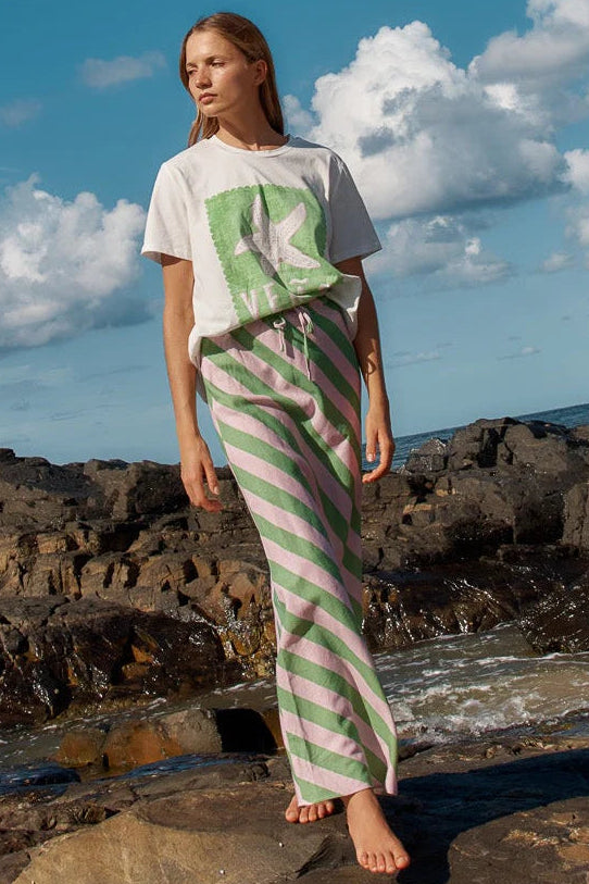 Woman wearing a white t-shirt with a green logo and striped pants standing on rocky terrain with ocean and sky in the background.