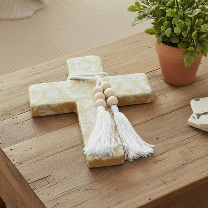A decorative wall cross featuring a mustard yellow and white floral and bird pattern, adorned with a white rope, wooden beads, and white tassels, shown against a plain white background.