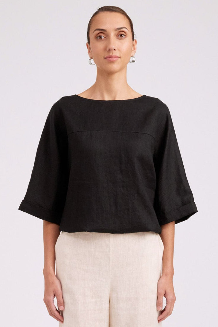 A model stands against a light gray studio background wearing a black short-sleeve top with a wide boat neckline and cream wide-leg trousers.