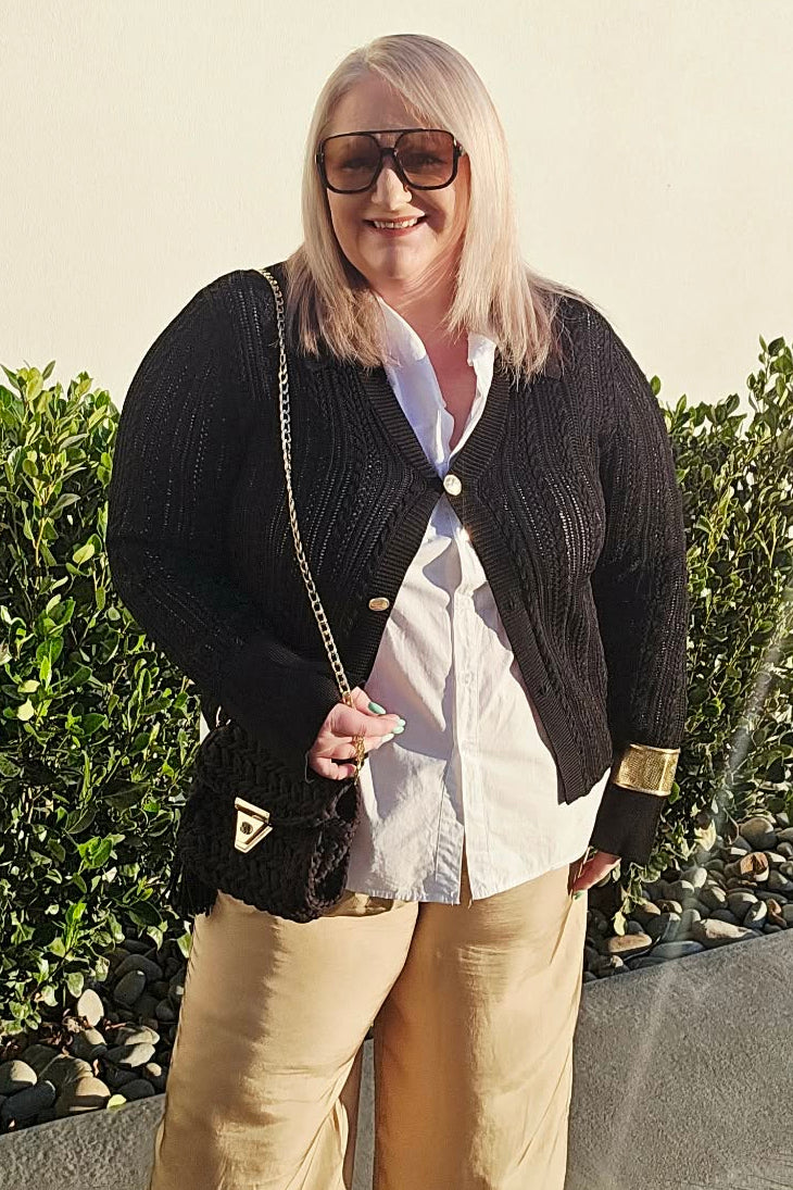 A woman posing outdoors wearing a black knit cardigan over a white button-up shirt, paired with wide-leg beige trousers and carrying a black shoulder bag.