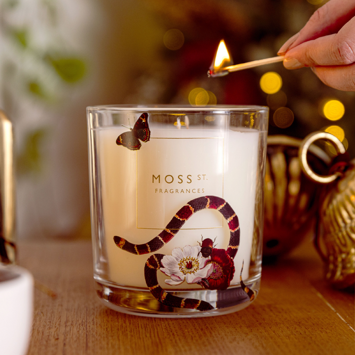 A glass jar candle with white wax and a printed snake, butterfly, and flower design sits on a wooden table. A hand holds a lit match near the candle, with a warm, softly blurred holiday background.