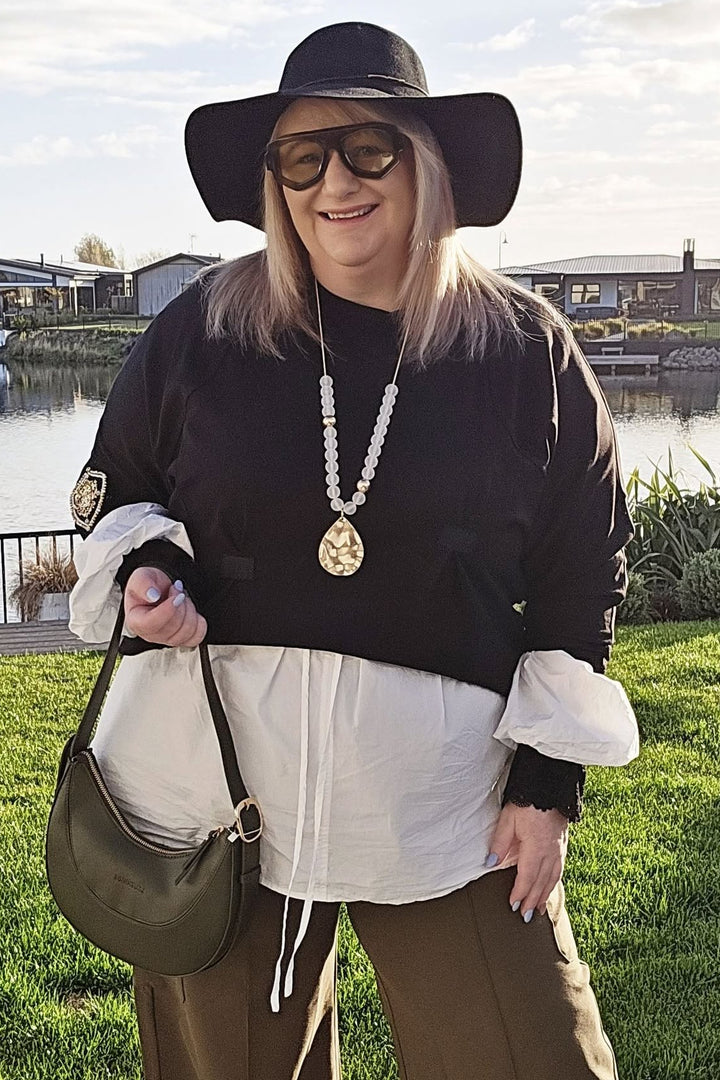 Woman standing outdoors by a waterfront, wearing a black top, white shirt, brown pants, and a wide-brimmed hat.
