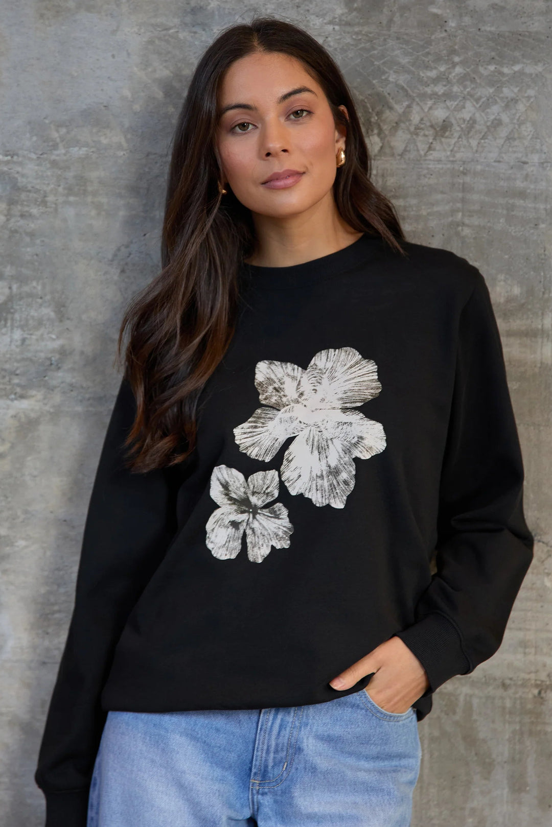 Woman wearing a black sweatshirt with white floral designs against a gray concrete wall.