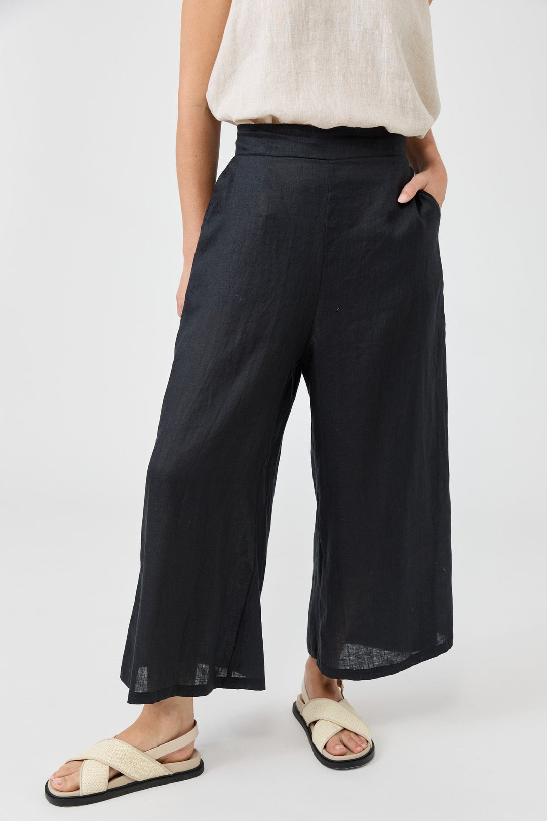 A model wearing black wide-leg cropped pants with side pockets, paired with a cream linen top and beige sandals, against a light gray studio background.