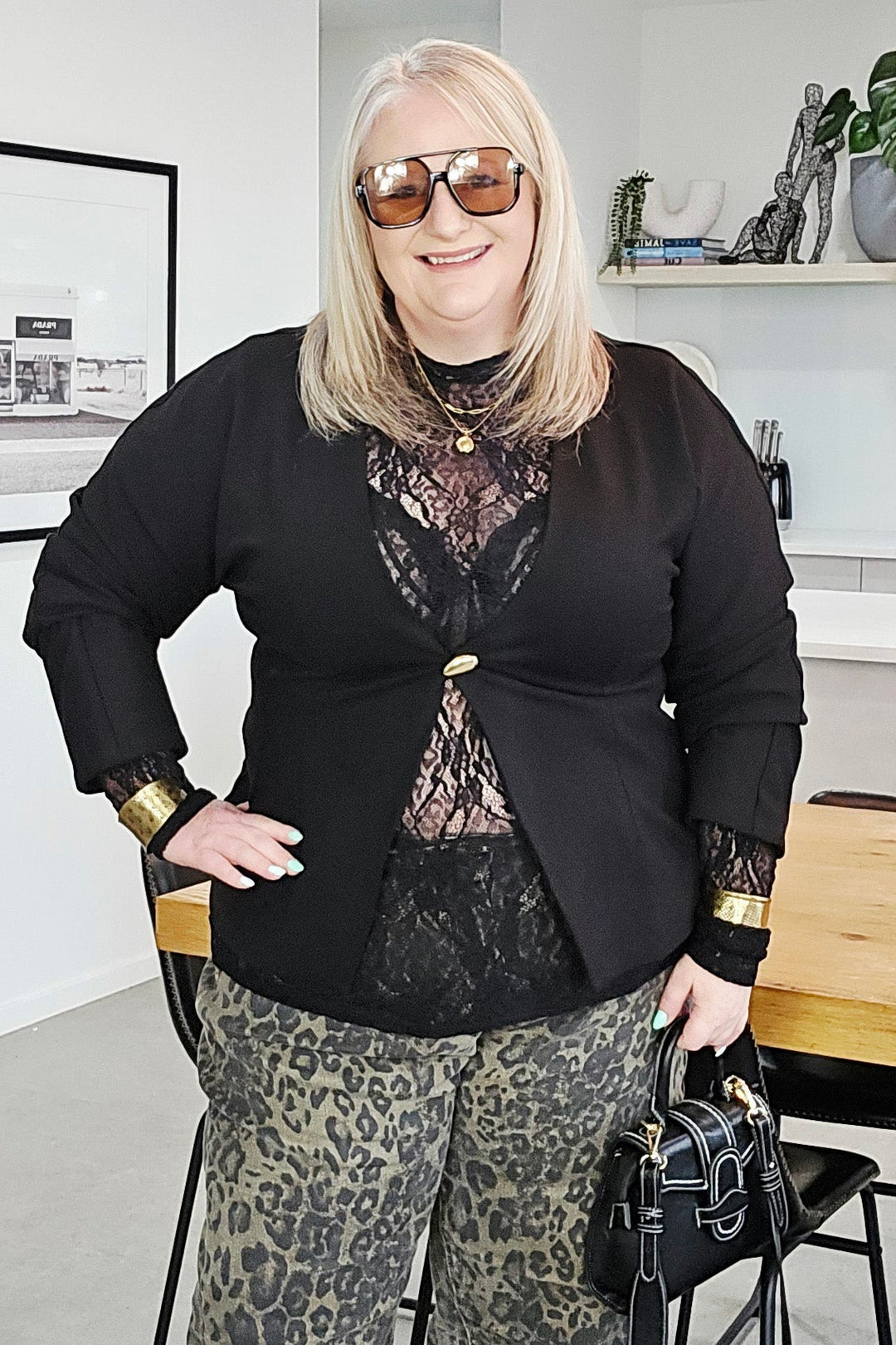A woman stands indoors modeling a black long-sleeve top with sheer lace detailing and wide-leg leopard-print pants. She is holding a small black handbag and wearing sunglasses in a modern room with a wooden table and shelves in the background.