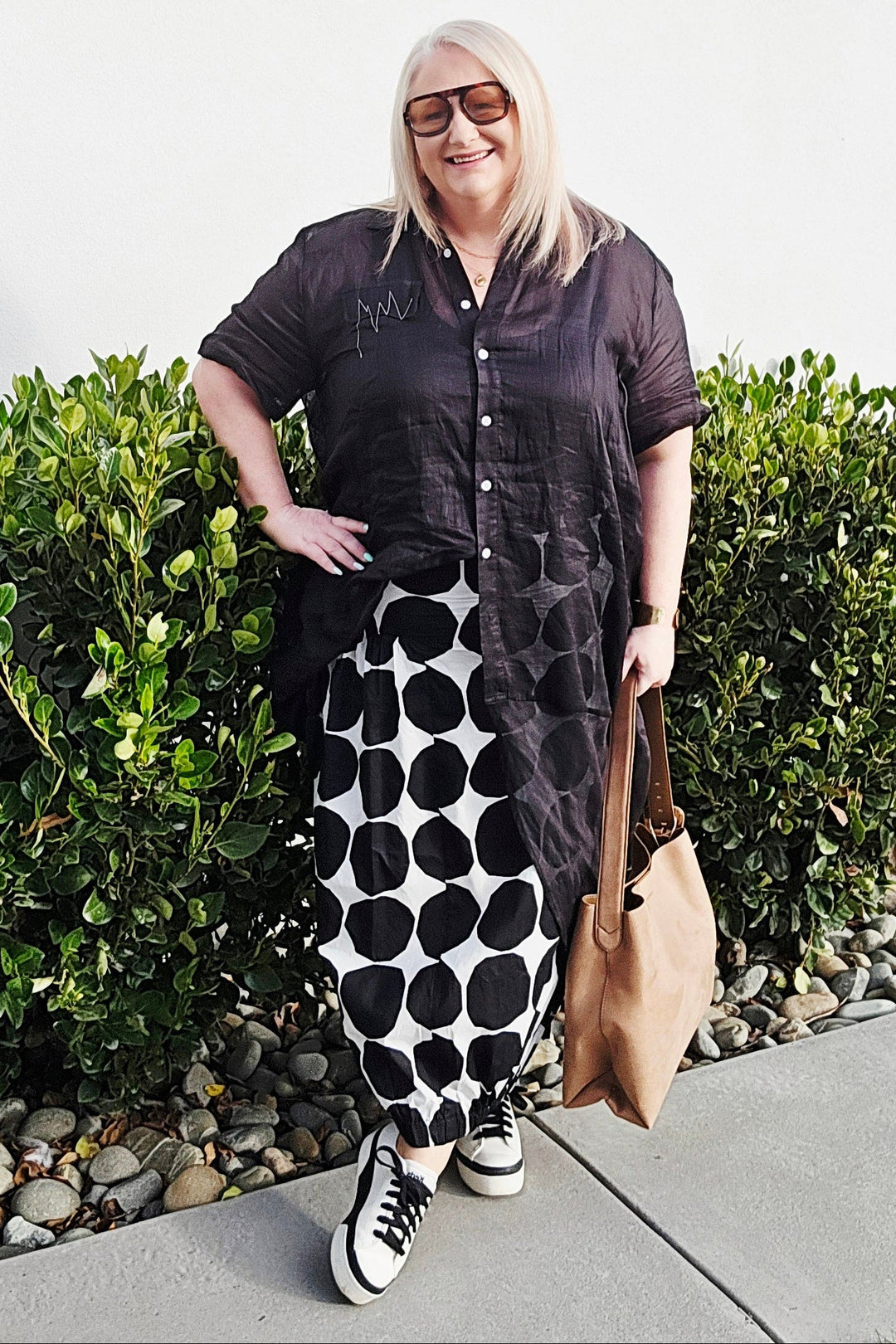 A woman posing outdoors in a black short-sleeve button-up shirt and a black-and-white polka dot maxi skirt, holding a tan tote bag.