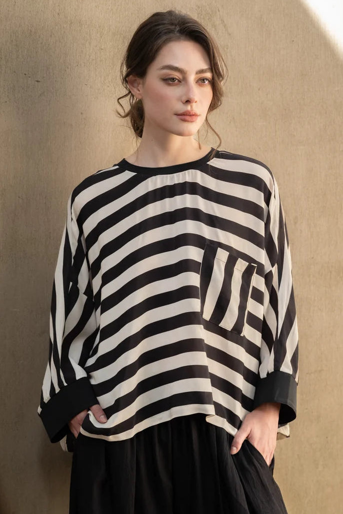 A woman wearing a black and off-white striped knit top with a chest pocket and black trousers, standing against a textured beige wall with warm sunlight and shadows.