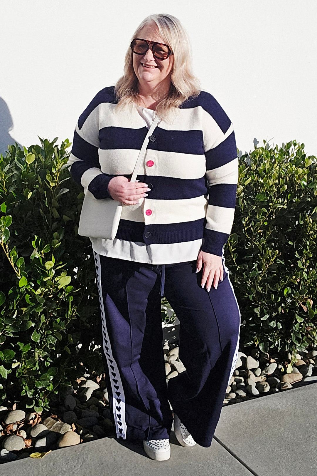 A woman wearing a navy and white horizontally striped cardigan, navy pants with white side stripes, and white sneakers, standing outdoors on a concrete path next to green bushes.