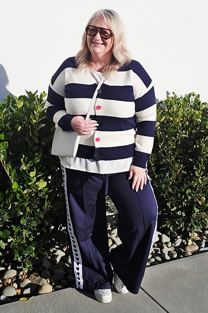 A woman wearing a navy and white horizontally striped cardigan, navy pants with white side stripes, and white sneakers, standing outdoors on a concrete path next to green bushes.