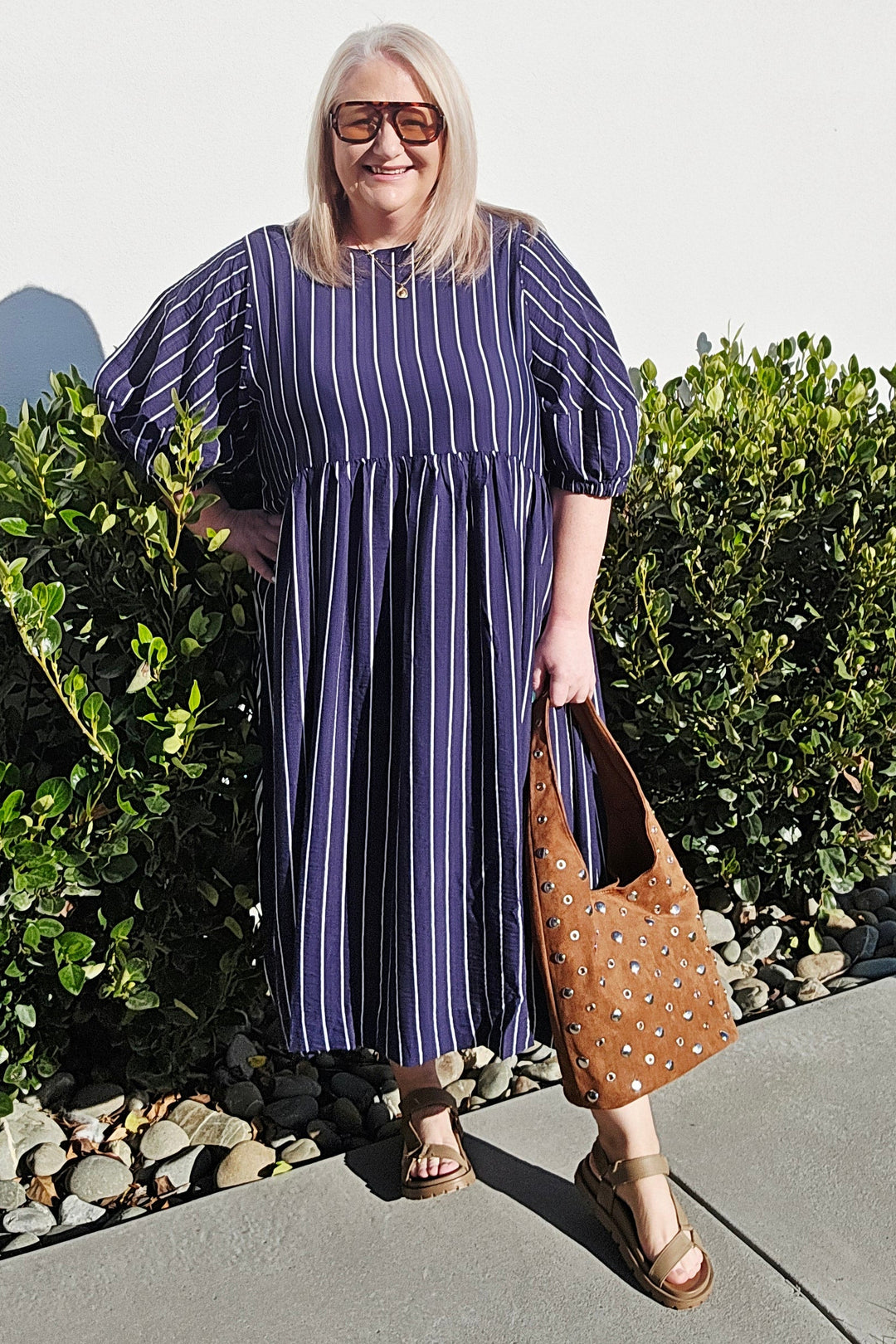 A woman wearing a blue and white vertically striped midi dress with puff sleeves, standing outdoors on a concrete path next to green bushes and holding a brown studded shoulder bag.