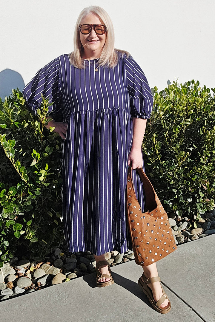 A woman wearing a blue and white vertically striped midi dress with puff sleeves, standing outdoors on a concrete path next to green bushes and holding a brown studded shoulder bag.