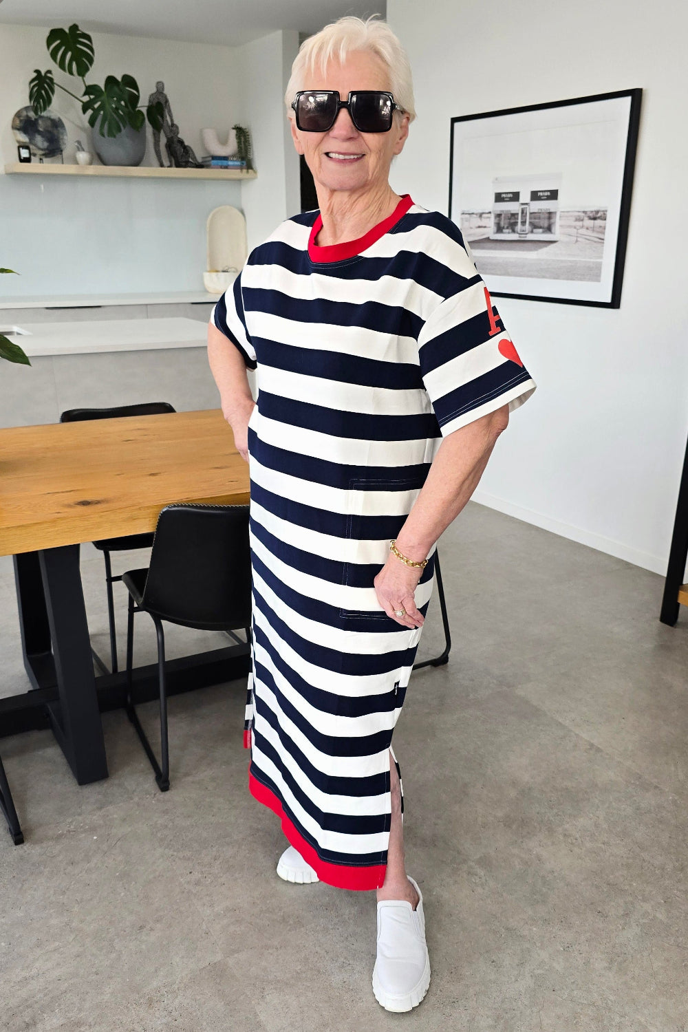 An older woman with short white hair and sunglasses stands in a modern room wearing a navy and white horizontally striped midi dress with red trim, white slip-on shoes, and a gold bracelet.