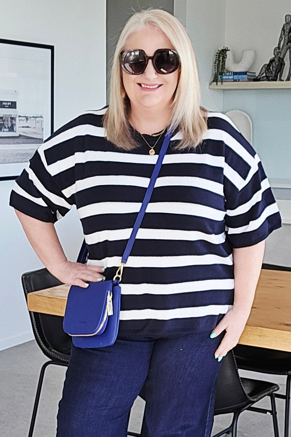 A woman stands in a modern home interior wearing a navy and white striped short-sleeve top, navy wide-leg pants, white sneakers, and carrying a blue crossbody bag.