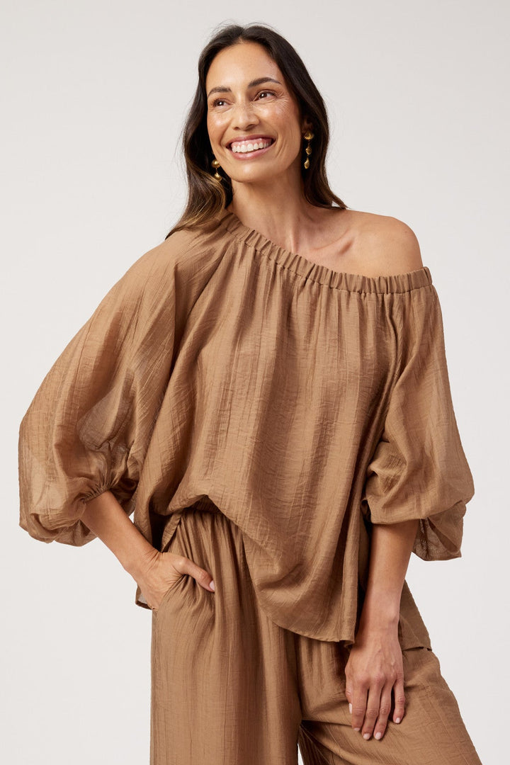 A woman modeling a two-piece brown set consisting of an off-the-shoulder, balloon-sleeve top and matching wide-leg pants, photographed against a plain light gray studio background.