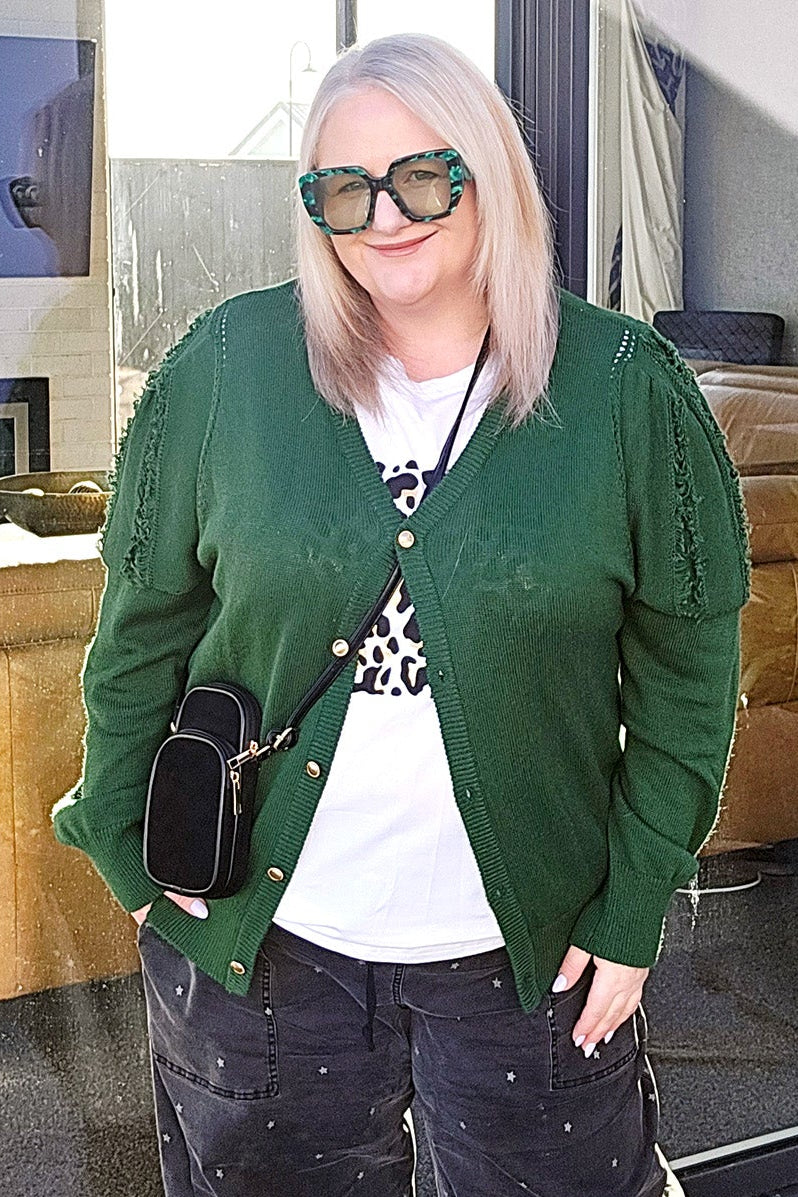 Person wearing a green cardigan, white shirt, and dark pants in front of a store window.