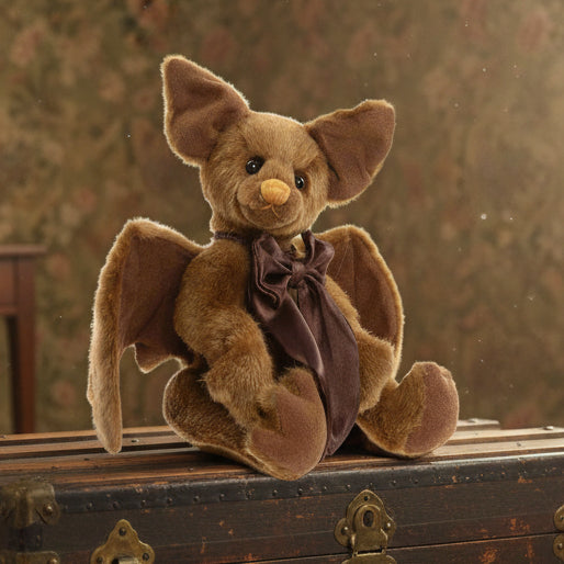 A plush toy resembling a bear with bat-like ears, named Pippy Strell, from the 2025 Charlie Bears Collection. It has a brown body and wings, with a darker brown bow accessory around its neck.