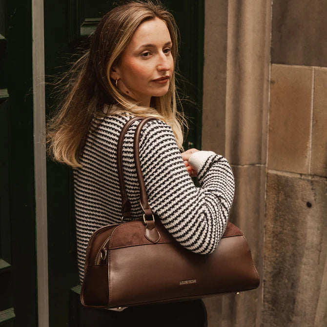 Woman holding a brown leather bag against a stone wall.
