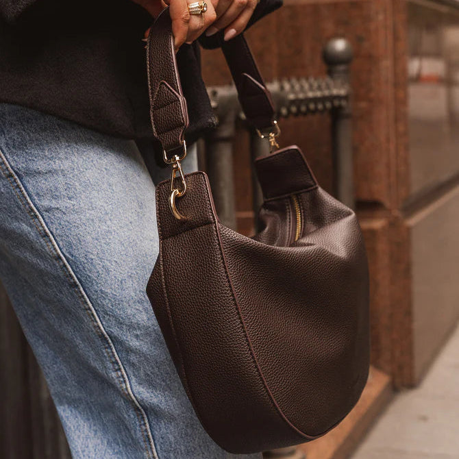 Brown leather handbag held by a person wearing a dark coat and blue jeans.