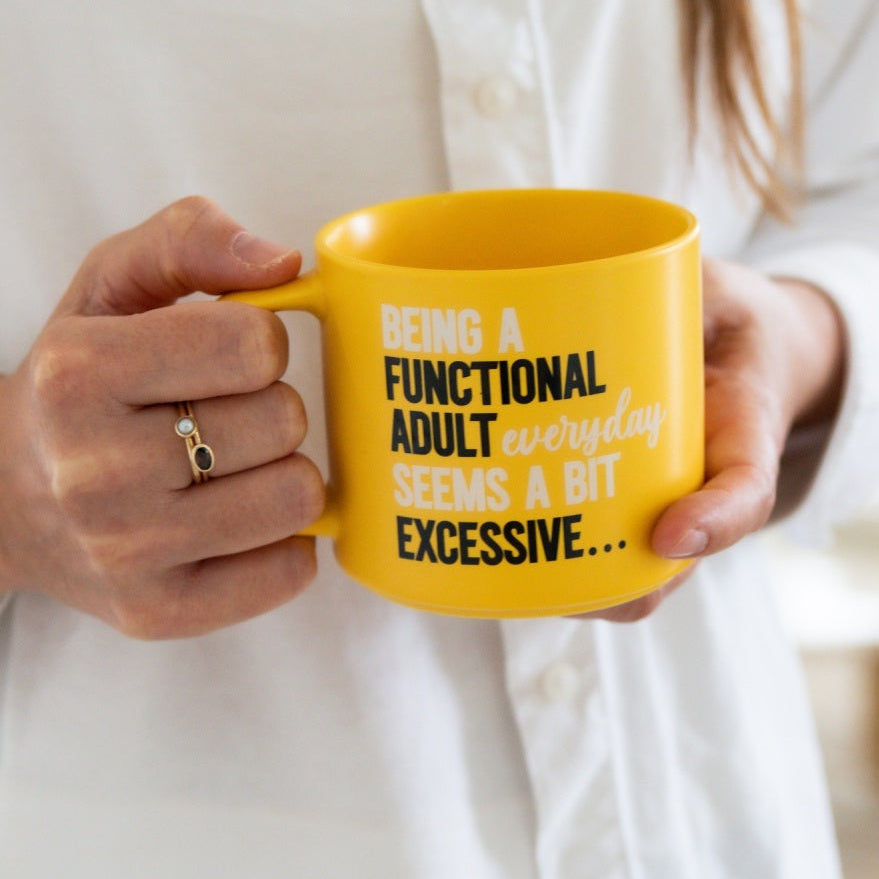 Yellow mug with text held by a person wearing a white shirt. MNug say Being a functional adult everyday seems a bit excessive
