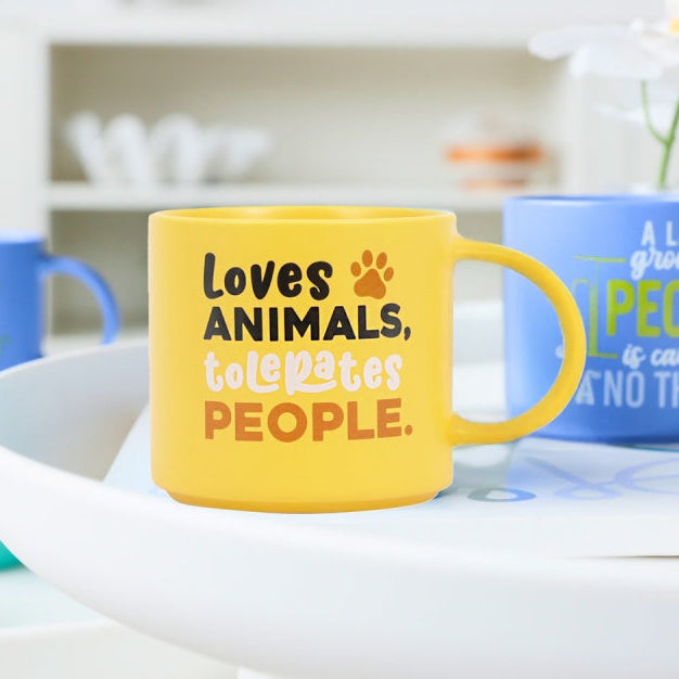 Yellow mug with text 'Loves animals, hates people' on a white surface with other mugs.