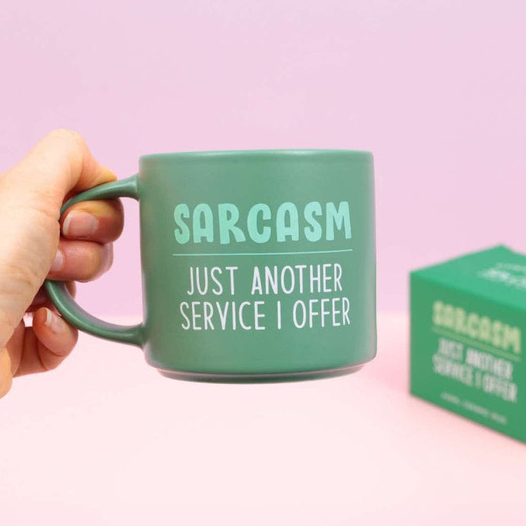 Green mug with 'Sarcasm Just Another Service I Offer' text, held by a hand, with a matching box on a pink background.