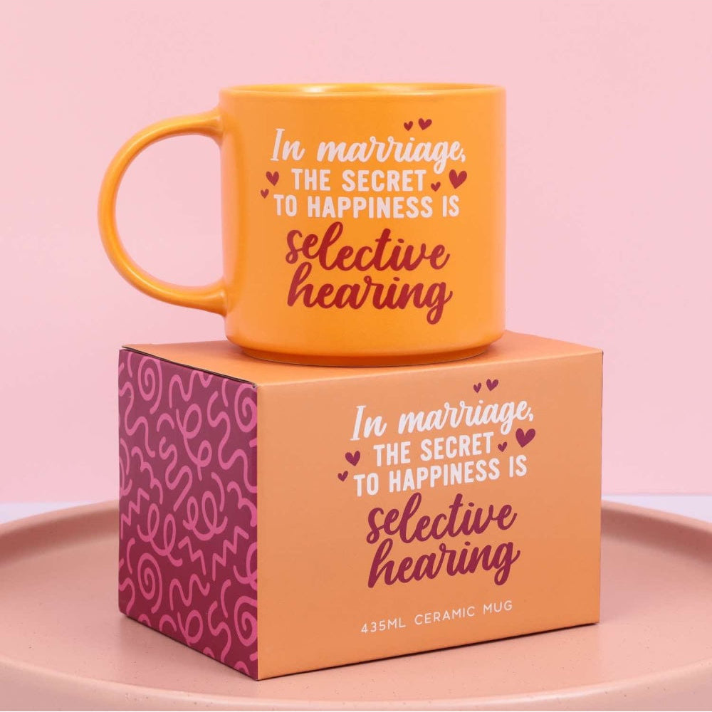 Orange mug with text 'In marriage, the secret to happiness is selective hearing' on a matching box against a pink background.
