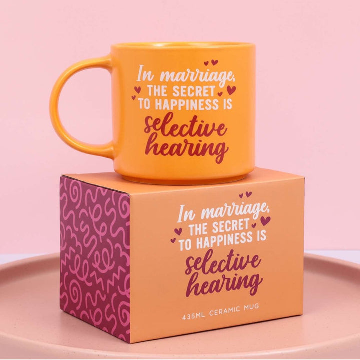 Orange mug with text 'In marriage, the secret to happiness is selective hearing' on a matching box against a pink background.