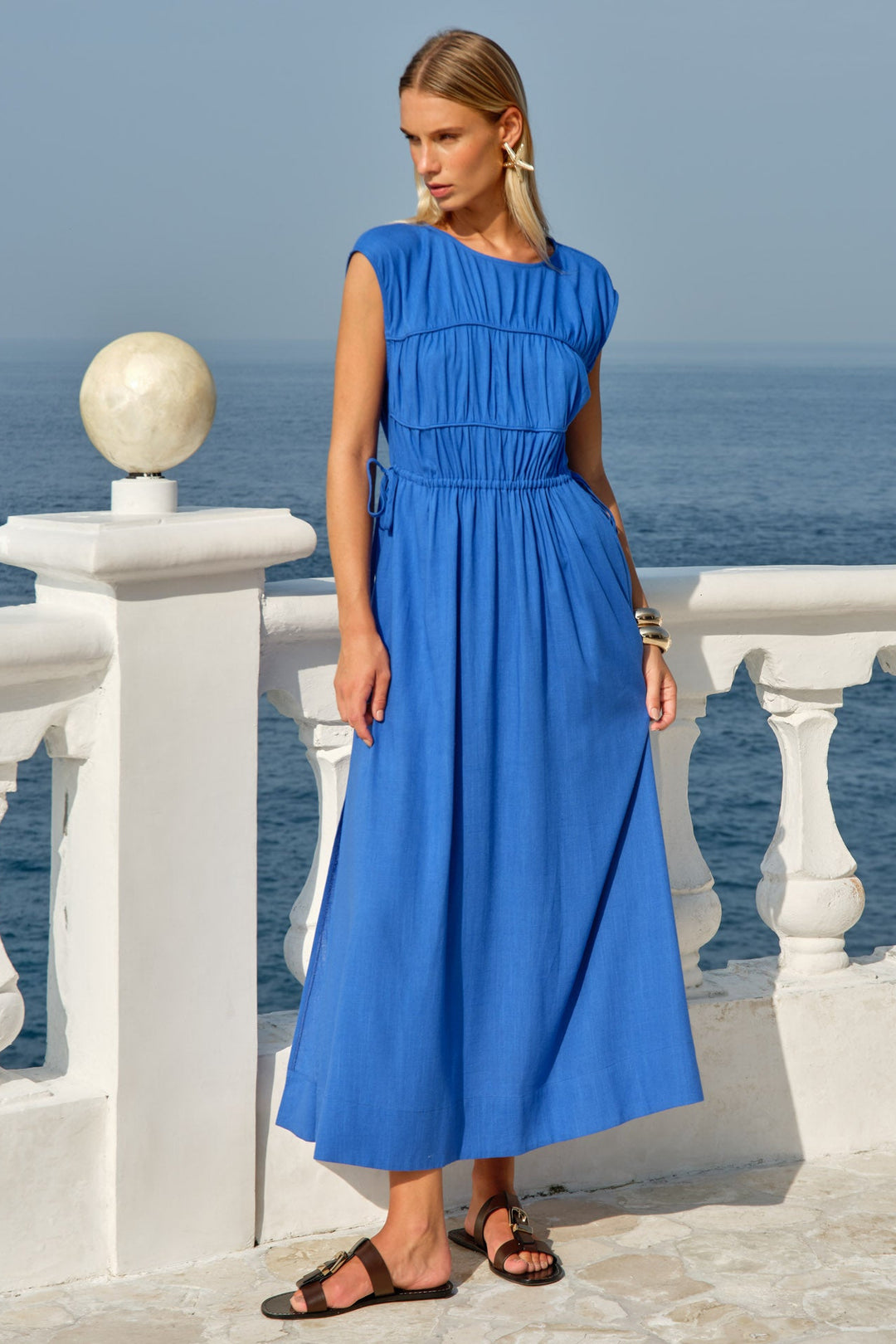 A woman wearing a bright blue sleeveless midi dress with a gathered waist and tie detail, standing on a white stone balcony overlooking the sea.
