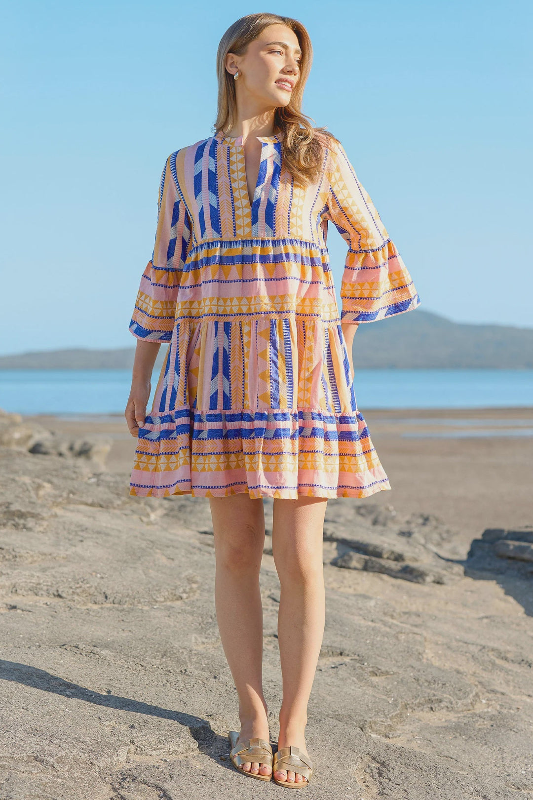 Woman in a colorful dress standing on a rocky beach with a clear blue sky.